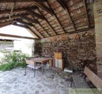 A stone stable with a wooden ceiling and simple furniture, surrounded by nature near a family house.