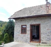 A stone cottage with a wooden roof in a mountainous area, with a garden and a view of the forests.