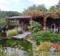 A cottage with a gazebo by the pond, surrounded by green nature and flowers.