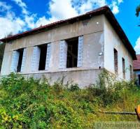 An abandoned family house with missing windows, surrounded by lush vegetation and a wooden fence.