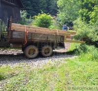 In the picture, there is a truck with wood by a cabin, surrounded by a forest and a path.