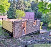 A wooden structure for a cabin is being built in the forest, surrounded by greenery and wooden cabins.