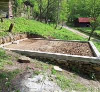 Preparing the foundation for a cabin with overgrown nature and tools next to an old house.