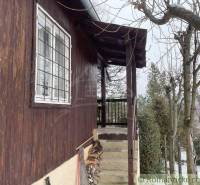 A wooden cabin with steps and firewood storage, with a snowy garden in the background.
