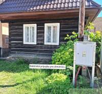 A log cabin with an electrical connection, a garden, and a family house in the background.
