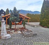A wooden gazebo in the garden of a family house with paving and a mountain backdrop.