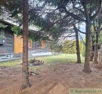 A wooden cottage surrounded by trees, a bench, a garden, and a family house in the background.