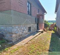 A family house with a stone foundation, next to a fence and a concrete road in a rural setting.