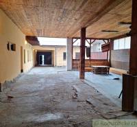 An elongated shelter with a wooden roof, an enclosed area, and a family house with a backyard.