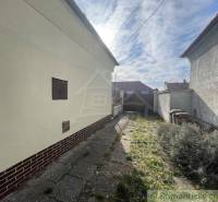A narrow courtyard of a family house with a concrete path leads to a double-winged gate.