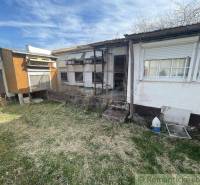 A yard with a neglected family house and an annex, surrounded by a lawn and a clear sky.