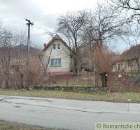 A family house surrounded by trees and bushes, a fence, a street, and a cloudy sky.