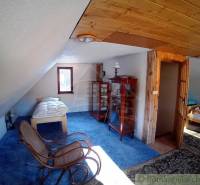 The interior of a cottage with wooden paneling, a bed, a display cabinet, and armchairs on a blue carpet.