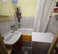 A bathroom in a 2-room apartment with a washing machine, bathtub, sink, and light tiles.