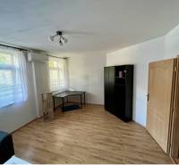 A room in a 2-room apartment with wooden flooring, air conditioning, furniture, and windows with blinds.