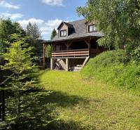 A cottage in Nová Bystrica with a garden and a forest backdrop under a blue sky.