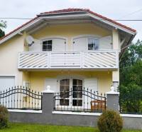 A family house in Dunasziget with white shutters and a balcony, surrounded by greenery.