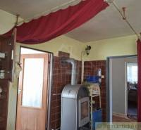 A room in a family house with a stove, curtains, and a shelf full of objects.