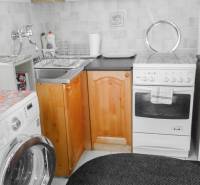 A kitchen in a 2-room apartment with a washing machine, stove, and wooden cabinets.
