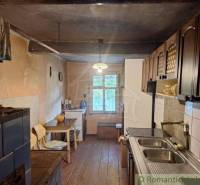 A kitchen in a cottage with a wooden floor, a tiled stove, and wooden cabinets.