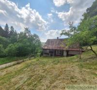 A log cabin in Zákopčie surrounded by a meadow and forest, under a blue sky with clouds.
