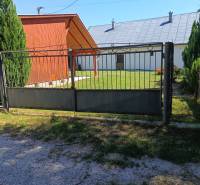 A gate and a wooden cottage in Tužina, surrounded by a lawn and coniferous trees.