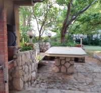 The courtyard of a family house in Vrsi with a stone table and a fireplace surrounded by greenery.