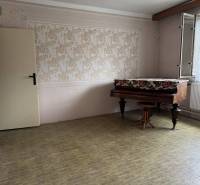 The interior of a family house with a piano, wallpaper, and an open window with a lace curtain.