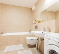 A bathroom with light tiles, a bathtub, a sink, and a washing machine in a studio apartment.