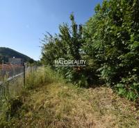 A sloped garden in Nová Baňa, enclosed by a fence, with a view of buildings and hills.