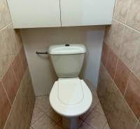 A toilet in a 4-room apartment with a cabinet above the tank and light tiles.