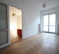 An empty room with parquet flooring leading to a room with a red couch in a 3-room apartment.
