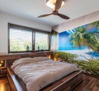 A bedroom in a family house with tropical wallpaper and a wooden ceiling fan.