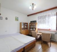 A room in a family house with a bed, a display cabinet, a table, and a plant by the window.
