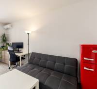 A one-room apartment with a sofa, a red refrigerator, and a desk by the open balcony.