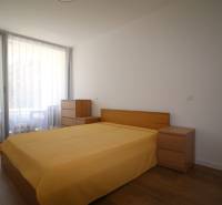 A bedroom with a bed and wooden furniture in a two-room apartment, light curtains letting in daylight.
