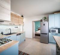 A kitchen in a 3-room apartment with wooden cabinets, flowers, and a view into the bedroom.