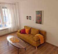 Living room of a two-room apartment with a yellow sofa, wooden table, and large window.