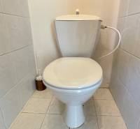 A toilet in a 3-room apartment with white tiles and a wooden panel on the wall.