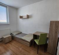 A simple room with a bed, a table, a chair, a nightstand, and a shelf in a building.