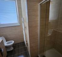 A bathroom in a building with a toilet, shower, and a window with blinds.