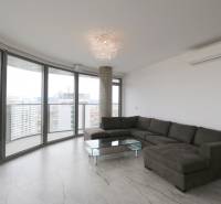 A bright living room in a three-room apartment with a corner sofa, glass walls, and a view of the city.