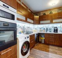 A kitchen in a family house with wooden cabinets, a stove, a washing machine, and decorative wallpaper.