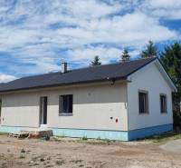 A family house in Kozárovce with a dark roof surrounded by greenery and construction materials.
