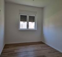 A room of a four-room apartment with wooden flooring, white walls, and a window.
