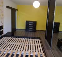 A bedroom in a 2-room apartment with a bed, two dressers, and a yellow wall.