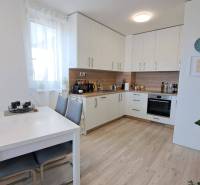 A kitchen in a 2-room apartment with a white kitchen unit, a dining table, and wooden flooring.