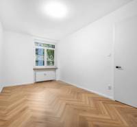 A room in a 3-room apartment with wooden flooring, white walls, and a window.