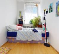 A room in a 3-room apartment with white walls, a bed, and a window with flowers.