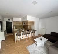 Living area of a 3-room apartment with an open kitchen, bar counter, and gray sofa.